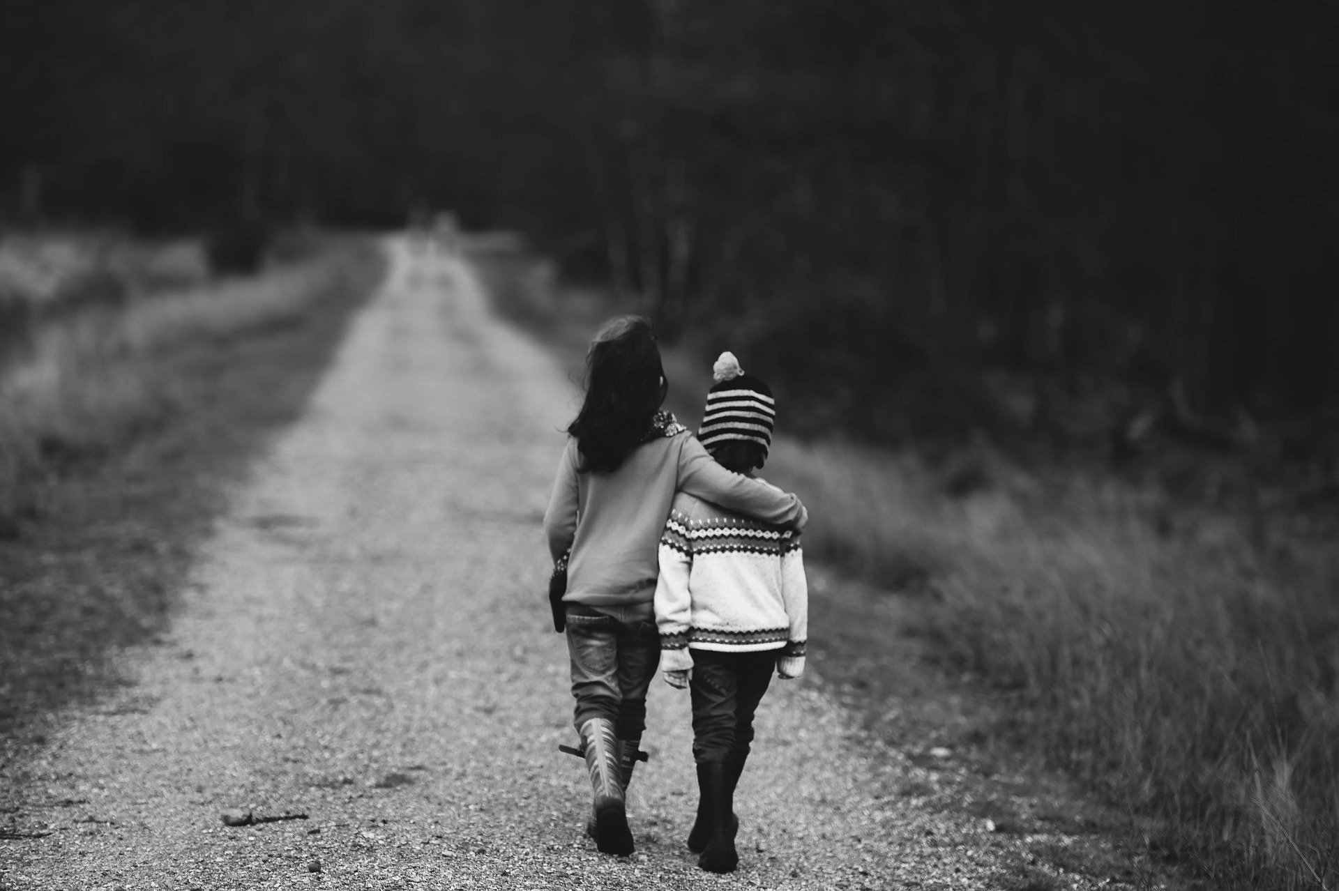 two kids, girl with arm around young boy, walking down a dirt road in the country