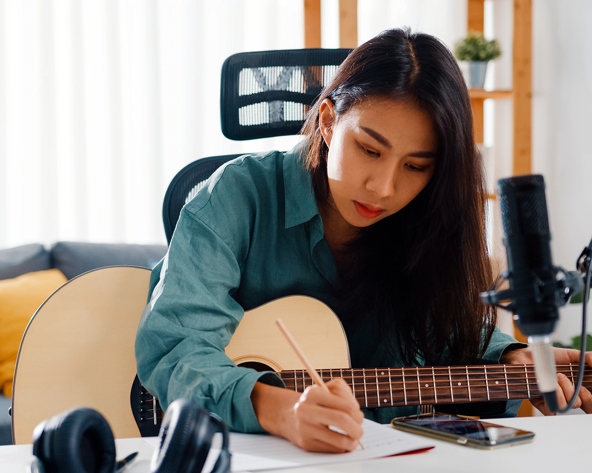 Happy asia woman songwriter play acoustic guitar listen song from smartphone think and write notes lyrics song in paper sit in living room at home studio. Music production at home concept.